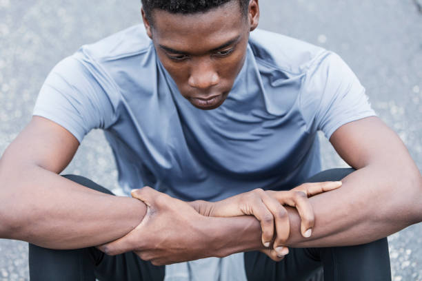Close-up of a serious young African American man sitting on the pavement. He is an athlete wearing sports clothing, in deep thought, looking down with a serious, sad expression.